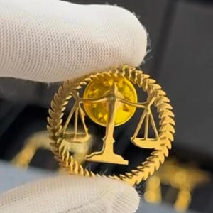 Könnte beinhalten: Ein goldfarbener runder Ansteckpin mit einem Waage-Symbol, umgeben von einem Lorbeerkranz-Design. Der Pin hat eine glänzende, reflektierende Oberfläche und ist wahrscheinlich aus Metall gefertigt. Der Hintergrund ist unscharf.