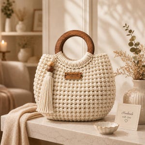 May include: A cream-colored crocheted handbag featuring a round dark wood handle, a matching cream tassel with wooden beads, and a rectangular brown leather-like tag centered on the front with the text Handmade with Love and a heart symbol.