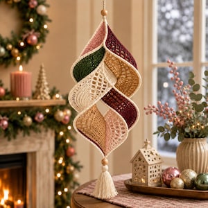 May include: A hanging spiral ornament crafted from crocheted yarn in shades of dusty pink, deep burgundy, forest green, and golden tan. The structure features an intricate openwork pattern, a wooden bead at the top and bottom, and a cream-colored tassel.