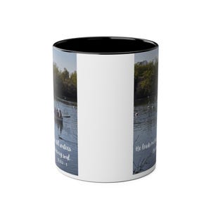 May include: A ceramic mug with a black interior and rim, featuring a wrap-around photographic print of a boat on a calm lake. The text on the mug reads, "He leads me beside still waters... He restores my soul... Ps 23:2-3" in a white script font.