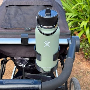 Op de afbeelding: Een saliegroene Hydro Flask waterfles met een zwarte dop en een flexibele draagbeugel, bevestigd in een zwarte plastic bekerhouder aan de duwstang van een zwarte kinderwagen. De fles is voorzien van een wit logo en de merknaam.
