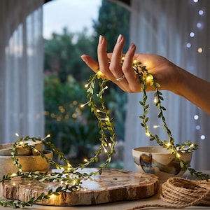 May include: A decorative string of artificial green leaves intertwined with small, warm white LED fairy lights. The flexible vine features numerous tiny, oval-shaped foliage pieces and glowing bulbs that create a soft, ambient illumination.
