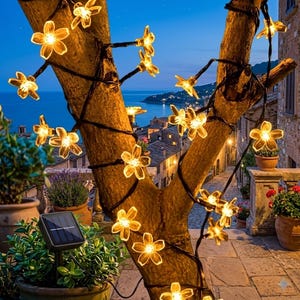 May include: A string of LED fairy lights shaped like five-petaled flowers wrapped around a tree trunk. The translucent yellow flower covers glow with warm light. A small black rectangular solar panel on a stake is positioned in a potted plant.