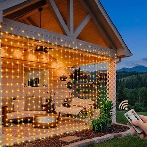 May include: A curtain of warm white LED string lights hanging from the roof of a covered porch, creating a grid pattern of illumination. A small handheld remote control with multiple buttons is shown in the foreground to operate the light display.