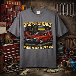 以下が含まれることがあります： 「DAD'S GARAGE」の文字と赤いクラシックカーのグラフィックがプリントされたグレーのTシャツ。「WHERE MONEY DISAPPEARS」の文字が車の下にあります。工具のあるガレージの背景にTシャツが置かれています。