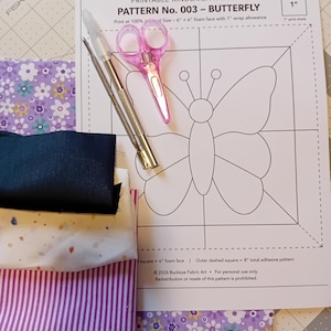 May include: A craft kit featuring a printed paper pattern titled BUCKEYE FABRIC ART PATTERN No. 003 - BUTTERFLY, a pair of pink scissors, a metal craft tool, and several fabric swatches including purple floral, solid navy, and white speckled.