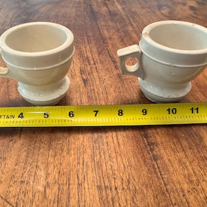 Puede incluir: Dos tazas de cerámica beige con asas, cada una con una base redondeada y un cuerpo cilíndrico. Una cinta métrica amarilla se coloca junto a las tazas, mostrando medidas en centímetros.