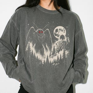 May include: A charcoal gray long-sleeve sweatshirt featuring a graphic print of a winged creature with glowing red eyes standing in a forest under a full moon. The word Pepper is printed vertically in the bottom left corner of the image.