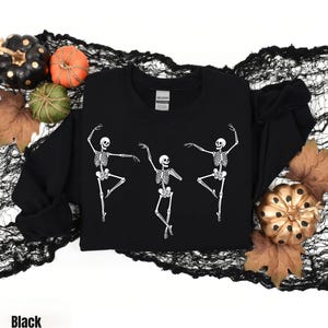 May include: A black long-sleeve sweatshirt featuring a white graphic print of three skeletons in various dancing poses. The garment has a crew neckline and a Gildan brand label at the inner collar. The design is centered on the front of the shirt.