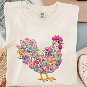 May include: An ivory-colored t-shirt featuring a central graphic of a chicken composed of various floral-patterned fabric patches in shades of pink, blue, and yellow. The chicken has a bright pink comb and wattle, a yellow beak, and yellow feet.