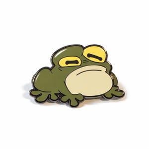 May include: A metal enamel pin shaped like a frog with olive green skin, a pale cream-colored belly, and large, sleepy yellow eyes. The design features bold black outlines and a smooth, glossy finish, capturing the frog in a low, resting pose.
