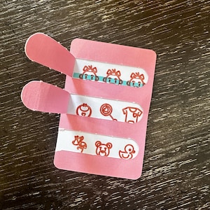 Puede incluir: Una tarjeta rosa con tres tiras blancas, cada una con pequeñas ilustraciones rojas. La tira superior tiene caras de mono, la del medio un caramelo y una camiseta, y la inferior una flor, un oso y un pato. La tarjeta tiene dos solapas rosas.