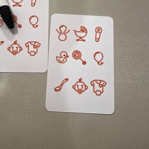 May include: Two white rectangular cards with rounded corners, each displaying red line drawings of baby-related items. The drawings include a baby carriage, safety pin, balloon, baby, onesie, duck, rattle, and spoon. A black marker is partially visible.