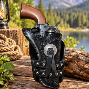 May include: A black leather holster with intricate floral tooling and silver-toned metal studs, featuring a central silver concho with a leather tie. A single-action revolver with a dark metal frame and a smooth brown wooden grip rests inside.