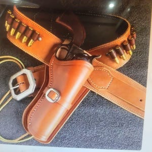Op de afbeelding: Een bruin leren pistoolholster- en riemset met een zilveren gesp. De riem heeft lussen met goudkleurige kogels. Het holster is ontworpen om een zwarte revolver te bevatten. De set wordt gepresenteerd op een donkergrijze ondergrond.