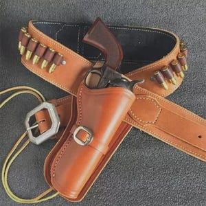 May include: A tan leather gun holster with a matching belt and bullet loops. A revolver is in the holster, and the belt has a silver buckle. The belt has a black interior.