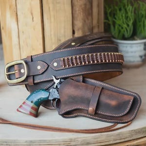 Può includere: Una cintura porta pistola in pelle marrone scuro con fibbia in ottone e una fila di passanti per proiettili. Un revolver con l'impugnatura dipinta con il motivo della bandiera americana in rosso, bianco e blu con stelle bianche è inserito.