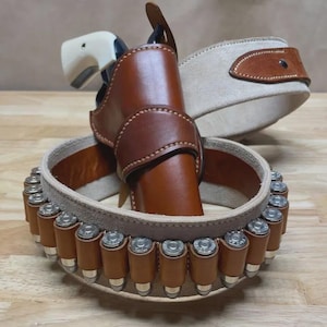 May include: A brown leather holster with a cream-coloured handle revolver. A matching brown leather belt with a cream-coloured suede lining and a cartridge belt with brown leather casings and silver bullets.