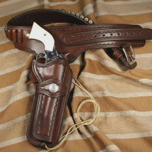 Könnte beinhalten: Ein braunes Lederholster mit passendem Gürtel, verziert mit Ziernähten, silberfarbenen Metallbeschlägen und einem silbernen Concho. Ein Revolver mit weißem Griff steckt im Holster, das mit einer dünnen, hellen Kordel versehen ist.
