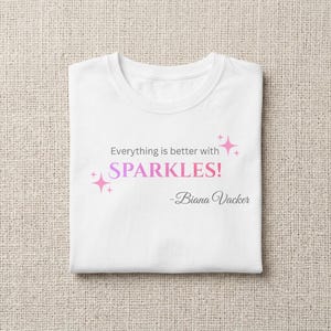 May include: White t-shirt with the text "Everything is better with SPARKLES!" in pink and purple, with a signature below. The shirt has a crew neck and is folded neatly.