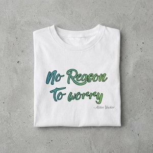 Op de afbeelding: Wit T-shirt met de tekst "No Reason To Worry" in blauw en groen script. De naam van de auteur, Alden Vacker, staat eronder. Het shirt is netjes op een getextureerd oppervlak gevouwen.