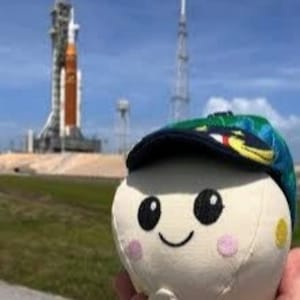 K&ouml;nnte beinhalten: Ein Pl&uuml;schtier mit einem l&auml;chelnden Gesicht, das eine blau-gr&uuml;ne Kappe tr&auml;gt. Der Hintergrund zeigt eine Raketenstartrampe und einen blauen Himmel. Das Spielzeug hat schwarze Augen, rosa Wangen und einen gelben Fleck.