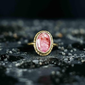 Könnte beinhalten: Ein goldener Ring mit einem ovalen, rosa Edelstein. Der Edelstein hat ein strukturiertes Aussehen mit roten und weißen Farbtönen. Die Fassung des Rings ist helleres Gold, und das Band ist dunkleres Gold.