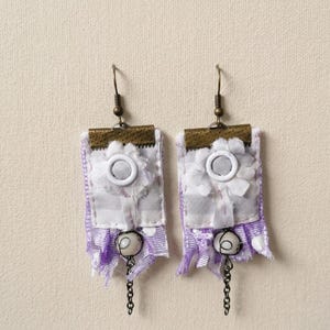 Puede incluir: Un par de pendientes hechos a mano. Cada pendiente presenta una base rectangular de tela con una flor blanca y un botón blanco en el centro. La base está adornada con cinta morada y una cadena colgante con una cuenta blanca.