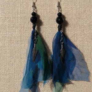 Può includere: Un paio di orecchini fatti a mano con perline nere, ganci argentati e tessuto blu e verde fluente. Ogni orecchino ha un piccolo ciondolo a forma di piuma argentata, creando un look bohémien. Un accessorio unico e accattivante.