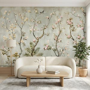 May include: A living room scene featuring a cream-colored, curved sofa, a light wood coffee table, and a gray rug. The backdrop is a mural with a floral design of flowering trees in soft pinks and yellows, with birds. A floor lamp and potted plant complete the decor.