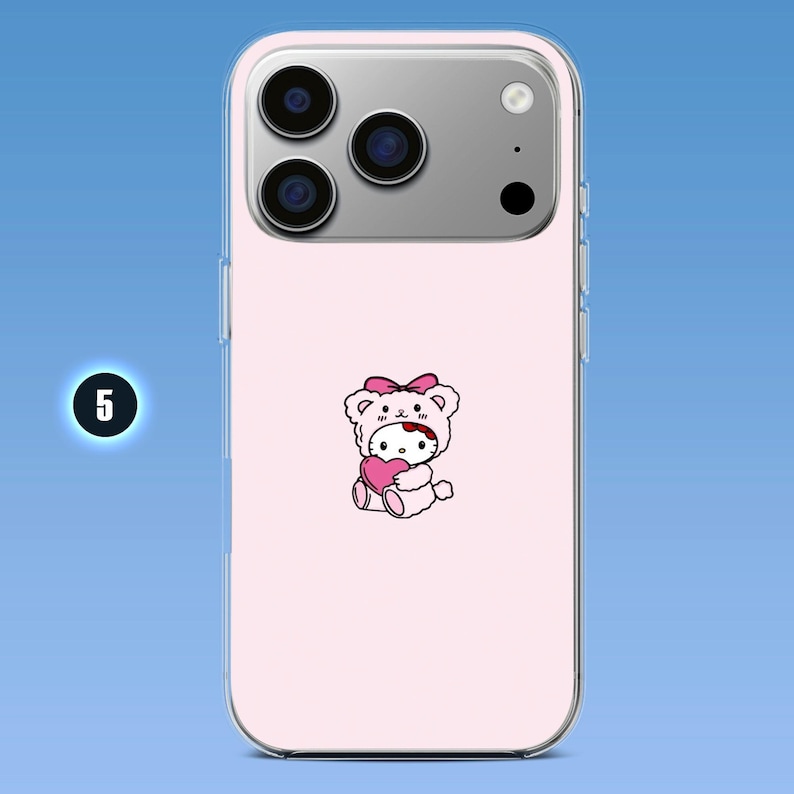 Puede incluir: Una funda de smartphone rosa claro con un gr&aacute;fico central de un personaje de gato blanco que lleva una capucha de oso rosa con un lazo, sosteniendo un coraz&oacute;n rosa. La funda tiene un borde transparente y una abertura para la c&aacute;mara triple.