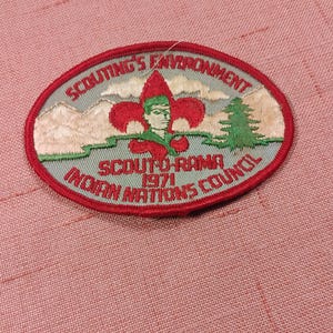 May include: An oval embroidered patch with a red border. The center features a red fleur-de-lis, a green pine tree, and mountains against a light blue sky. Text reads: SCOUTING'S ENVIRONMENT, SCOUT-O-RAMA 1971, and INDIAN NATIONS COUNCIL.
