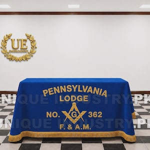 May include: A rectangular blue tablecloth featuring gold embroidered text that reads PENNSYLVANIA LODGE NO. 362 F. & A.M. with a central gold square and compass emblem containing the letter G. The tablecloth is finished with a gold fringe along the bottom.