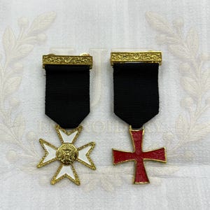 Puede incluir: Dos medallas suspendidas de cintas negras con barras doradas ornamentadas en la parte superior. La medalla de la izquierda es una cruz de esmalte blanco con un emblema central dorado. La de la derecha es una cruz de esmalte rojo con bordes dorados.