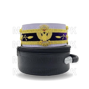 Puede incluir: Un sombrero ceremonial con bordados dorados, una banda de terciopelo púrpura y un emblema de águila. El sombrero descansa sobre un estuche de transporte negro con asa. El sombrero tiene el texto "REUM DELICIUM".