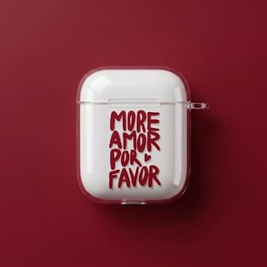 Op de afbeelding: Een witte AirPod-case met een doorzichtige beschermhoes. De case heeft de tekst "MORE AMOR POR FAVOR" in een vet, rood lettertype. De achtergrond is effen donkerrood.