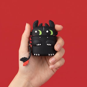 Pode incluir: Estojo de silicone preto para AirPod em forma de dragão de desenho animado, com olhos verdes e uma cauda vermelha e preta. O estojo tem um pequeno laço para prender a um chaveiro ou cordão. O rosto do dragão é detalhado com pontos brancos e uma boca sorridente.
