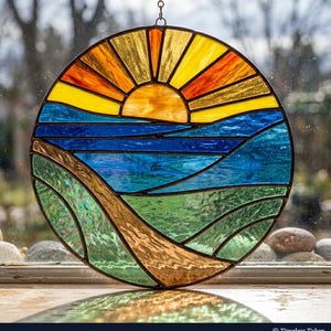May include: A round stained glass panel depicting a landscape scene. The design features a yellow and orange sun, blue water, and green and brown rolling hills. The panel is hanging from a chain.