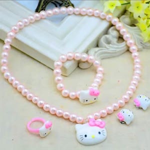 May include: A jewelry set consisting of a light pink pearl necklace and a matching bracelet, both featuring a white Hello Kitty charm with a pink bow. The set includes a matching pink ring and two white Hello Kitty stud earrings with pink bows.