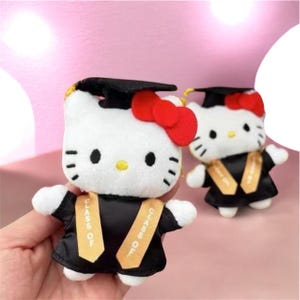 May include: Two plush Hello Kitty figures dressed in black graduation gowns and caps. Each figure features a bright red bow, black whiskers, and a yellow nose. The gowns include gold-colored stoles with the text "CLASS OF" printed on them.