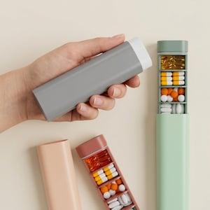 Puede incluir: Un pastillero rectangular gris con una tapa blanca se sostiene en una mano. Se muestran otros pastilleros en rosa y verde, cada uno con múltiples compartimentos llenos de pastillas de varios colores y formas. Los organizadores están diseñados para la medicación diaria.