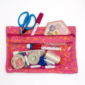 Könnte beinhalten: Eine pinkfarbene und orangefarbene Stofftasche mit Polka-Dots und einer transparenten Vorderseite, gefüllt mit Nähutensilien. Inhalt: blaue Schere, Marker, Faden und ein Stoffstück mit Blumenmuster. Die Tasche ist etwa 25 cm breit.