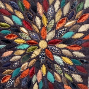 Può includere: Un'opera d'arte tessile quadrata con un motivo floreale centrale. Il fiore è composto da numerosi pezzi di tessuto colorati a forma di foglia in tonalità di rosso, arancione, blu, verde e oro, disposti su uno sfondo blu scuro. I petali sono in velluto e raso.