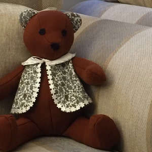 Può includere: Un orsacchiotto marrone con un gilet a fantasia bianca e marrone è seduto su un divano a righe.