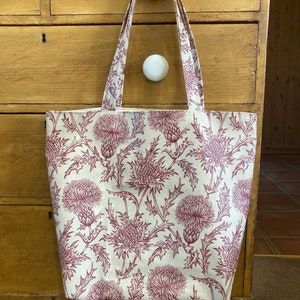 Puede incluir: Una bolsa de tela blanca con un estampado floral de cardos color burdeos. La bolsa tiene dos asas largas.