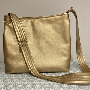 Puede incluir: Un bolso de hombro de cuero de color dorado con una correa ancha. El bolso tiene una forma rectangular simple y un acabado liso y brillante.