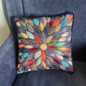May include: Square decorative pillow with a dark blue background and a colorful leaf-like pattern. The leaves are various colors including orange, teal, yellow, and red. The pillow has a dark blue border.