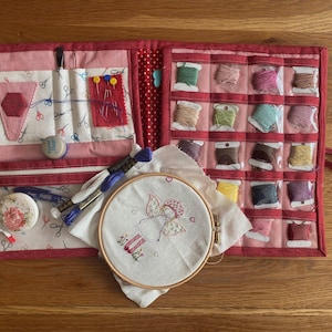 May include: Open red fabric sewing kit with various compartments. Includes spools of thread, embroidery floss, needles, pins, scissors, a tape measure, and an embroidery hoop with an angel design. The kit is open on a wooden surface.