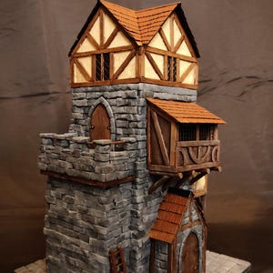 Puede incluir: Una torre de piedra en miniatura con una base de mampostería gris, una escalera de madera apoyada contra la pared, un techo de tejas marrones y un nivel superior de entramado de madera con paredes blancas. Incluye un balcón y puertas arqueadas.