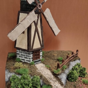 Puede incluir: Un modelo de molino de viento en miniatura con una torre beige de entramado de madera marrón oscuro, base de piedra gris y cuatro aspas claras. La estructura se asienta sobre una base de paisaje texturizada con camino de grava y arbustos.
